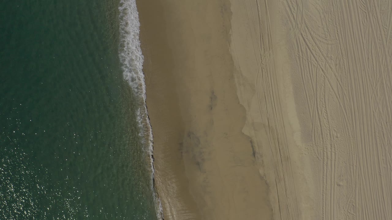 Overhead view of a Baja California Sur shoreline with green-blue ocean on the left, a narrow foam edge, tan sand with tire tracks on the right, and no visible people.