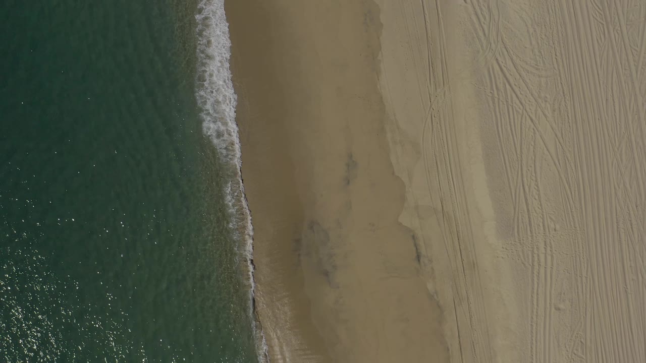 Drone-style overhead frame of the Baja California Sur coast, with rippled ocean, white wave foam, darker wet sand, patterned dry beach, and no visible people.