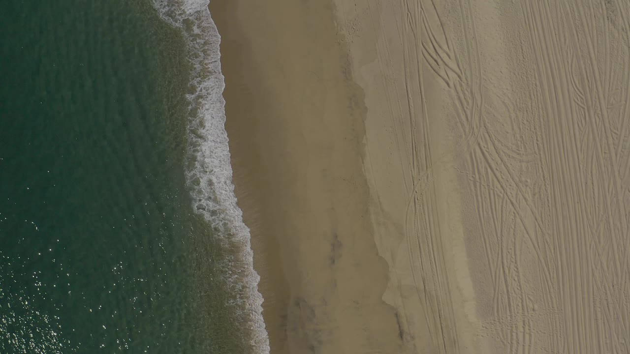 Aerial view along the Baja California Sur beach edge, showing foamy water curving into damp sand and multiple vehicle tracks across the dry sand, with no visible people.