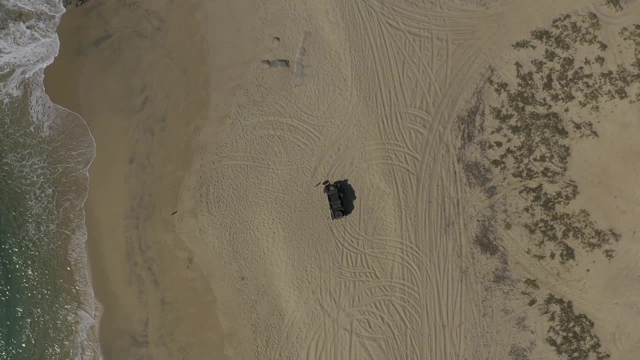 Top-down Baja California Sur shoreline frame with a strip of ocean surf at left, dense tire marks through the sand, vegetation on the right, a dark vehicle slightly right of center, and no visible people.