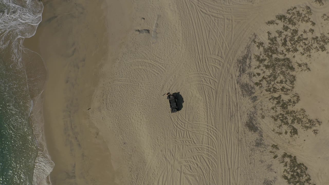 Overhead Baja California Sur beach frame with foamy water entering from the left, broad tracked sand across the middle, darker vegetation patches to the right, a dark vehicle below center, and no visible people.