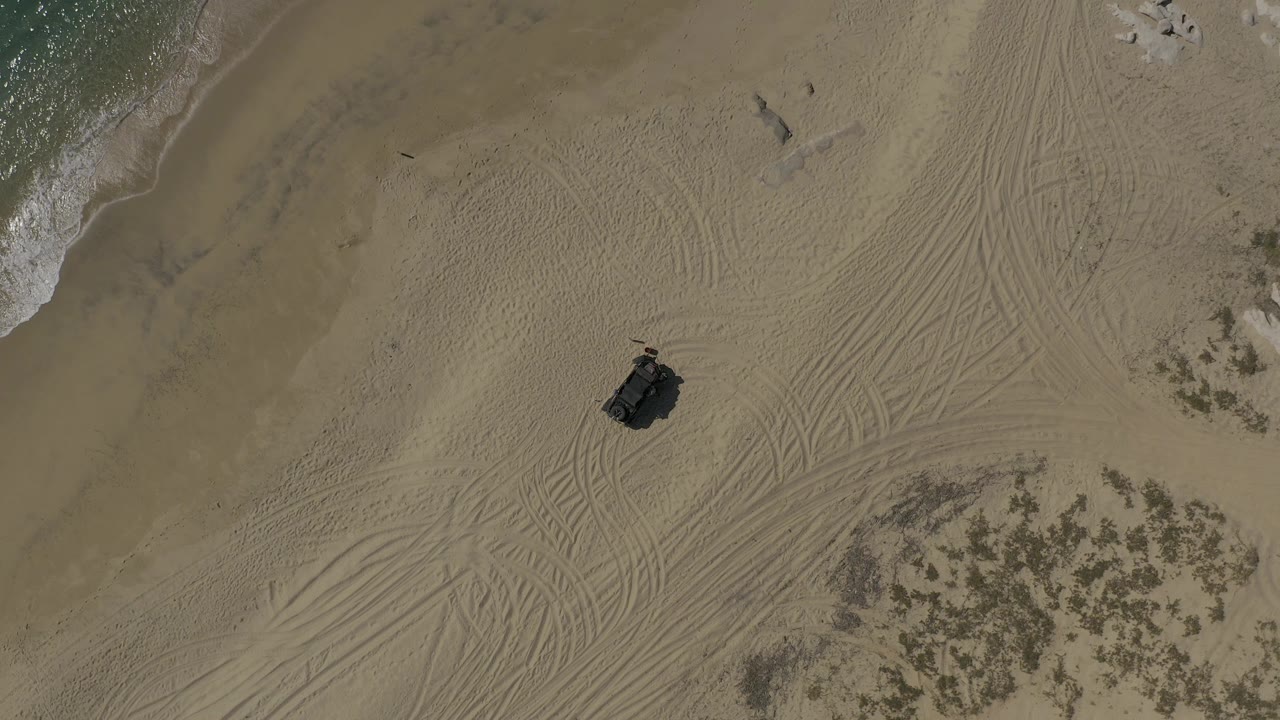 Aerial view over Baja California Sur sand with tire tracks crossing the frame, a dark off-road vehicle or tent-like object near center, scattered scrub, rocks, and surf along the upper edge, with no visible people.