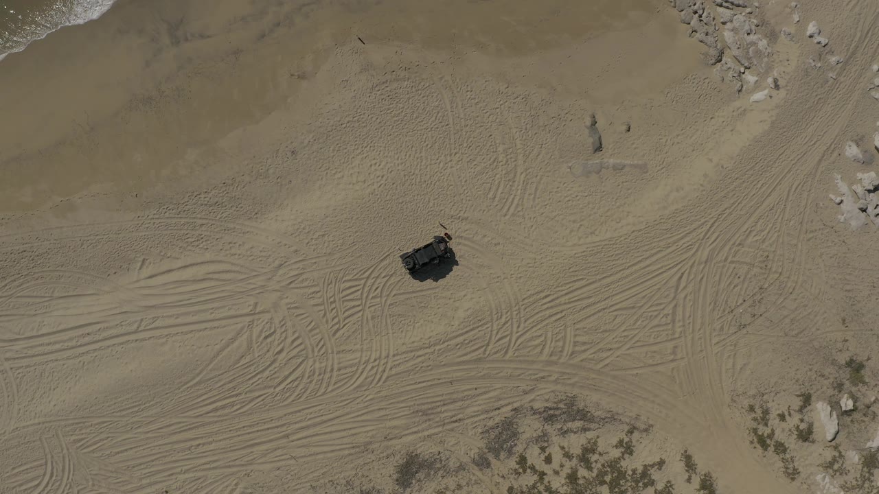 Aerial view over Baja California Sur coastal sand where a dark off-road vehicle or tent-like object sits on open tracked sand, with pale rocks near the upper right and no visible people.