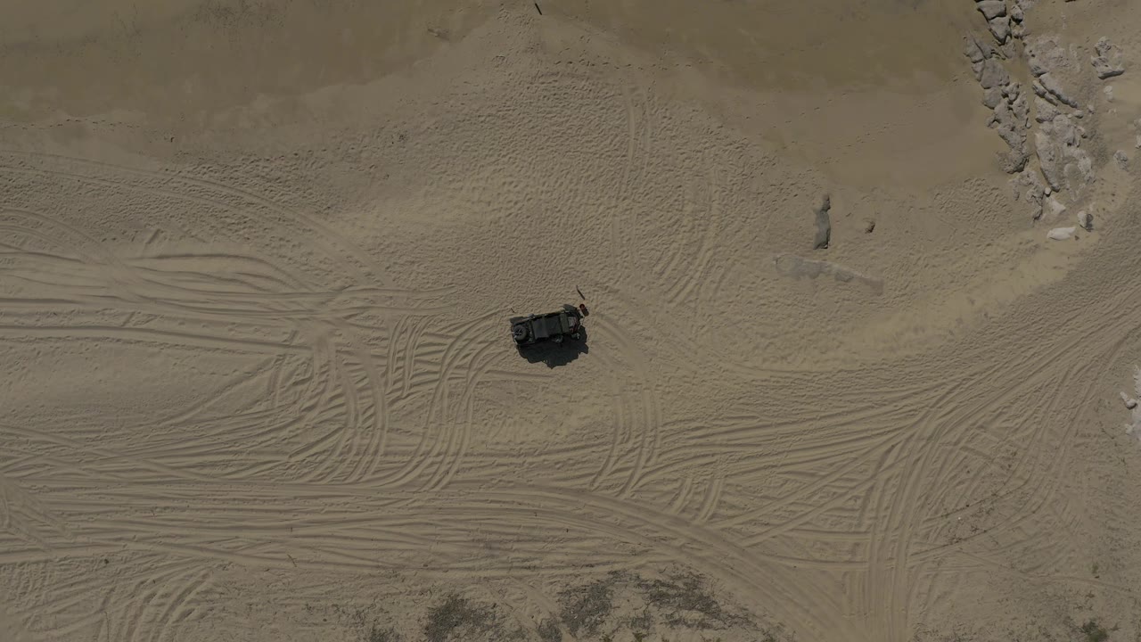 High aerial frame in Baja California Sur, Mexico, with the dark vehicle near the lower-left area, crisscrossing tire marks across the sand, pale rocks above, and no visible people.