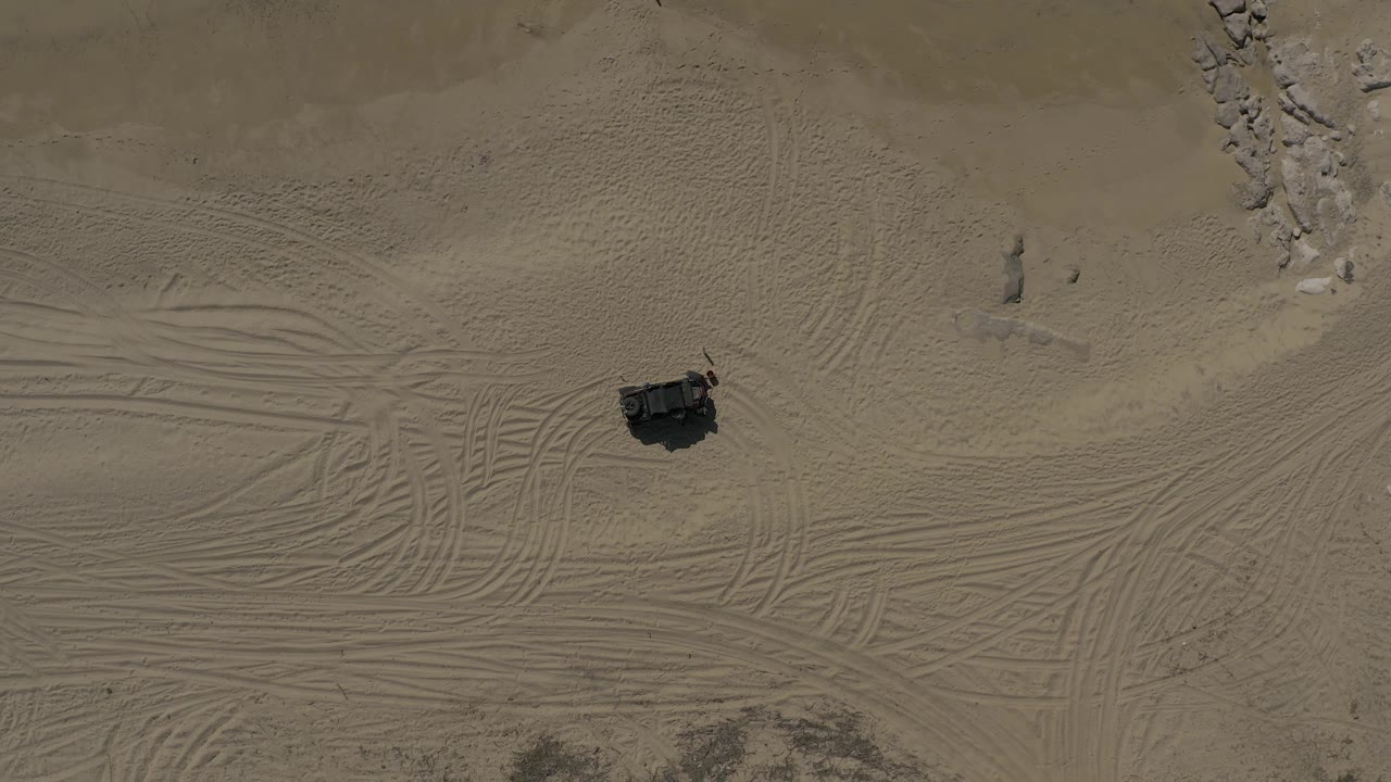 Overhead Baja California Sur desert-beach sand frame with the vehicle below center-left, dense tire-track patterns in the midground, pale rocks at the upper right, and no visible people.