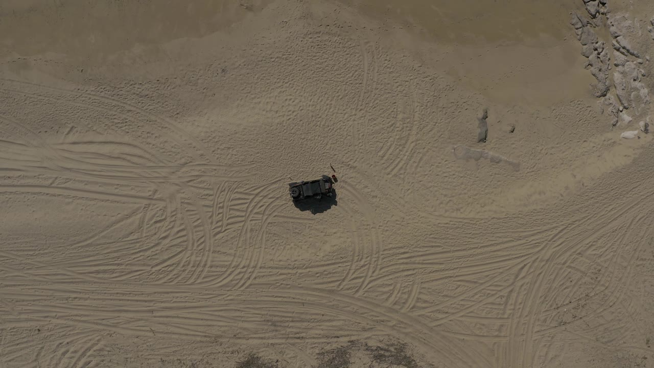 Aerial frame in Baja California Sur, Mexico, showing an off-road vehicle near the lower-left side, broad tire marks sweeping across tan sand, scattered rocks above, and no visible people.