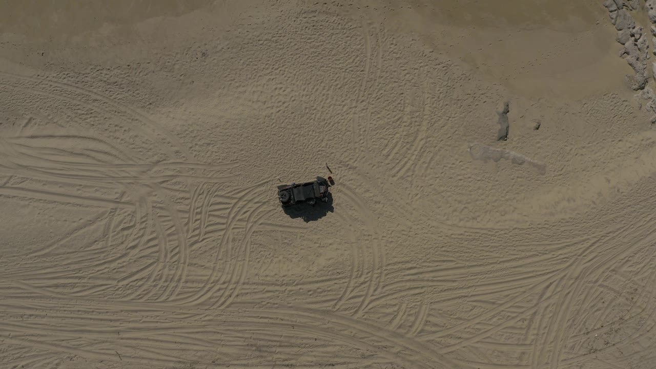 Straight-down aerial frame in Baja California Sur showing pale sand crossed by tire tracks, a dark vehicle near the lower left area, and no visible people.