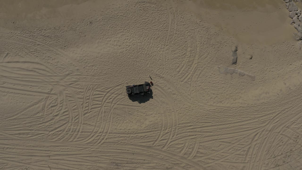 Straight-down aerial view in Baja California Sur with the dark vehicle near the lower center, crisscrossed tire tracks, faint sand ripples, and no visible people.