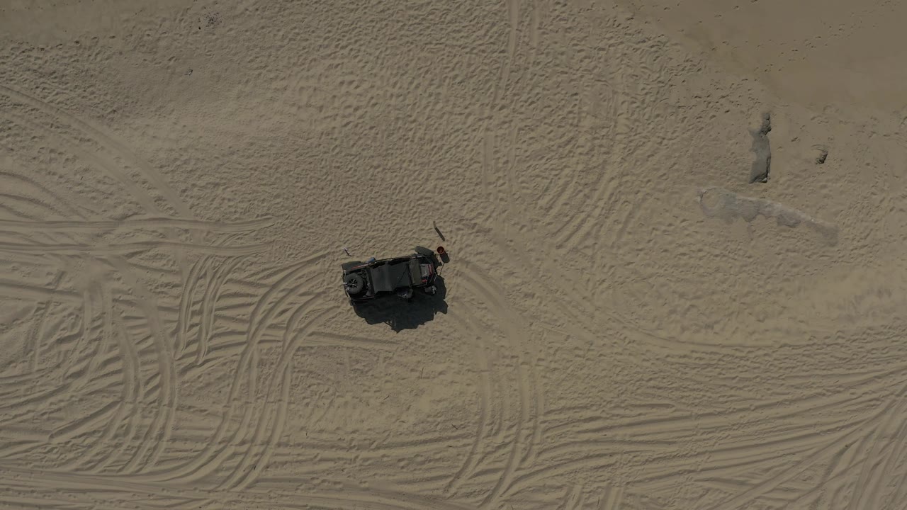 High aerial view in Baja California Sur Mexico with the dark object slightly more centered among curved tire tracks and low dune texture, with no visible people.