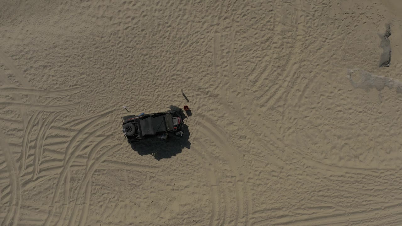 Overhead frame of a black off-road vehicle on pale sand in Baja California Sur, Mexico, with tire tracks beside it and no visible people.
