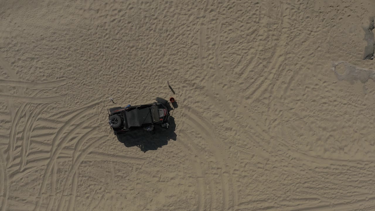 Overhead drone-style view of a dark vehicle on textured sand in Baja California Sur, Mexico, with track marks to one side and no visible people.