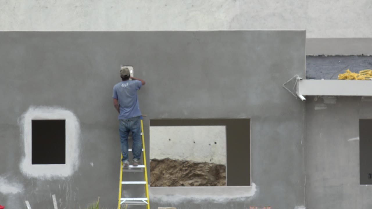 At 0:01, the worker faces the upper gray wall from the ladder, with the dark patched window to the left and a ledge with yellow material near the top right.