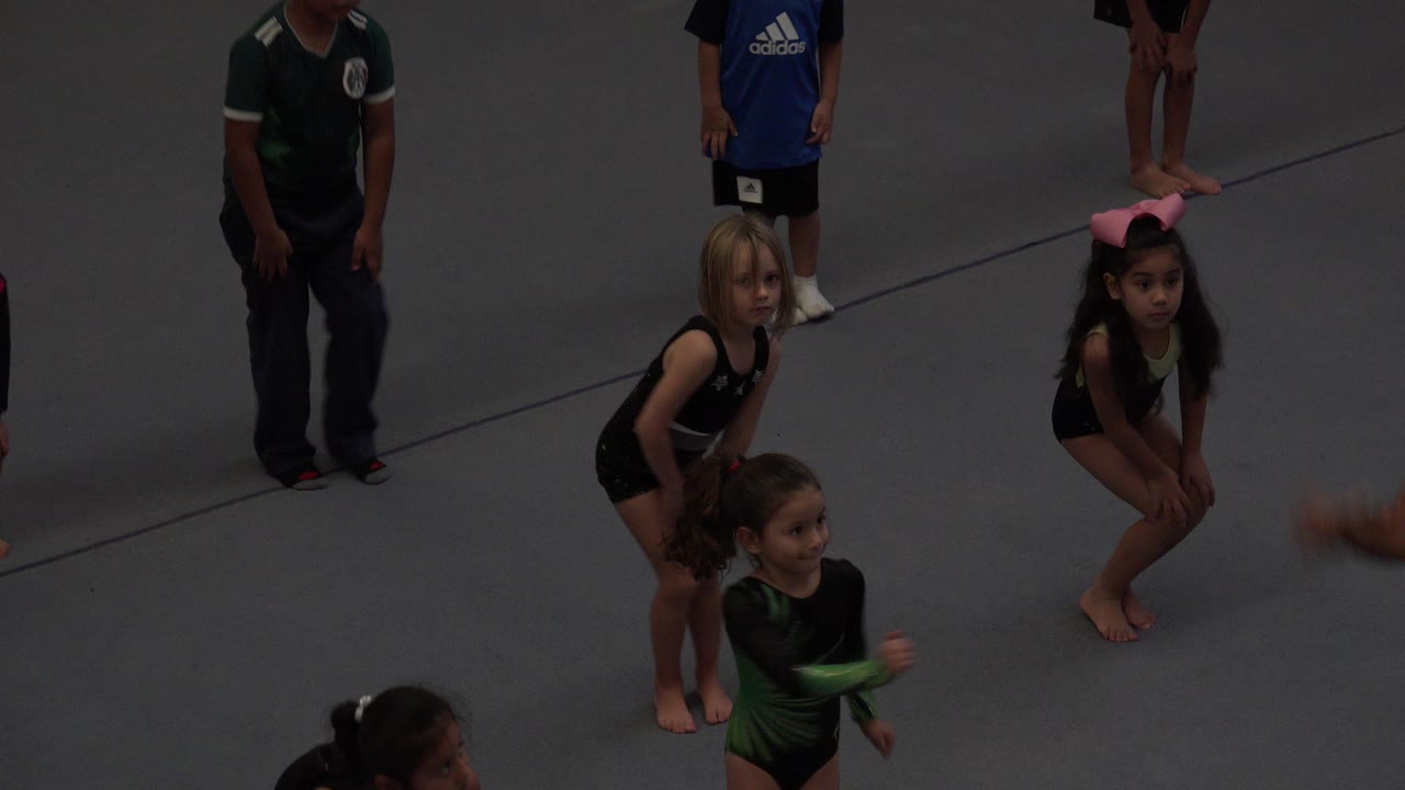 Children in gymnastics outfits stand on a gray mat in Cabo San Lucas, Mexico, with faces visible and an adult partially visible near the edge of the frame.