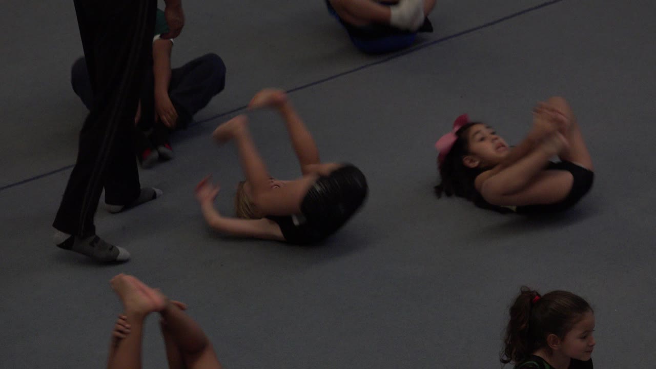 Children rolling on a gray padded floor as an adult stands nearby.