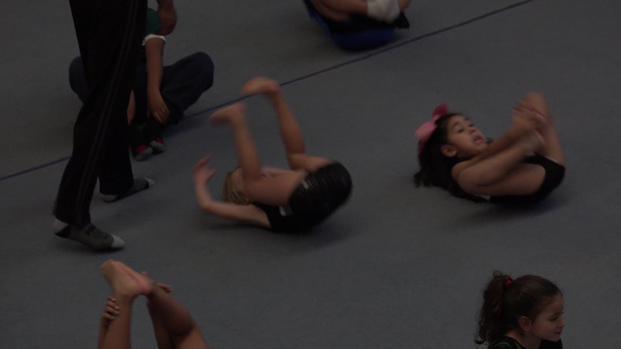 In Cabo San Lucas, Mexico, two children lie on their backs on the gray mat with knees raised while an instructor stands at the edge of the class.
