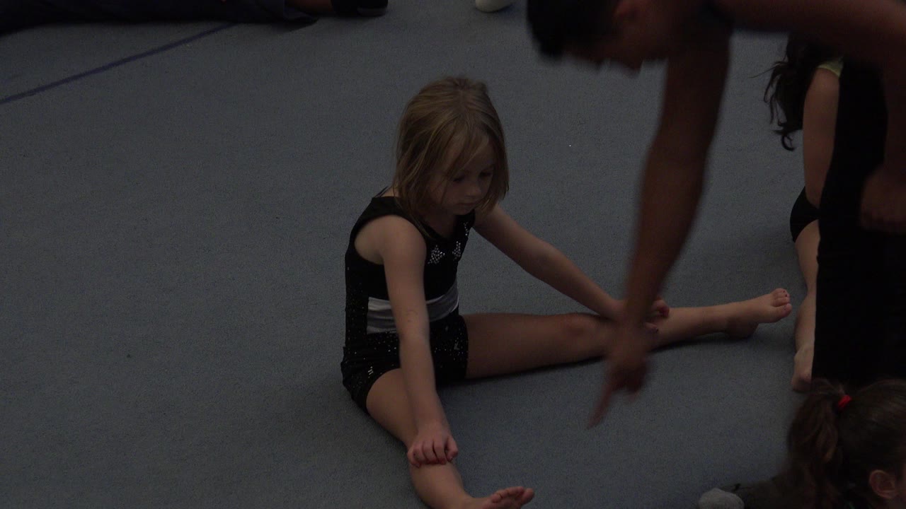 Young girl sitting in a split stretch on a gray gym floor with an adult instructor nearby.