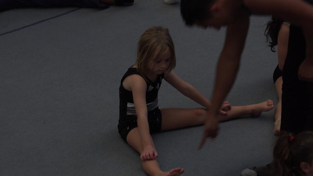 In Cabo San Lucas, Mexico, the instructor's arm points down near the seated girl's foot as another child sits partly behind her on the gray floor.