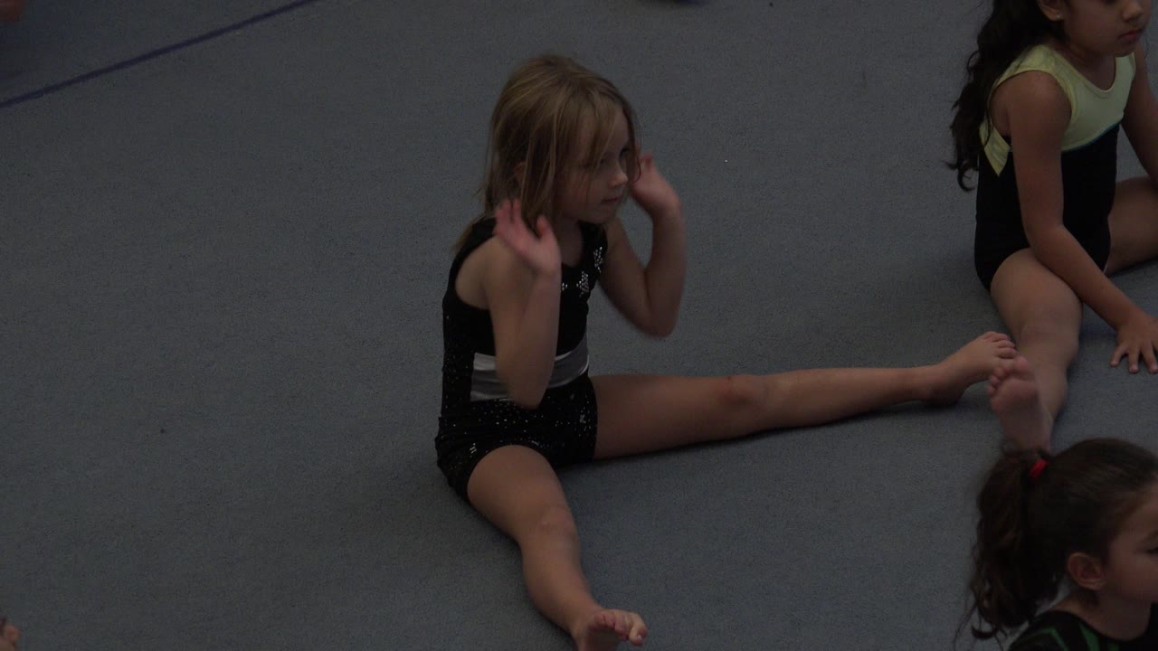 Young girl sitting in a split stretch on a gray mat with other children nearby.