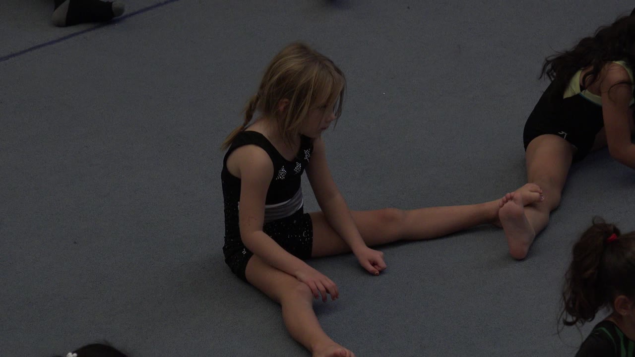 Two young girls sitting on a gray padded floor during class.