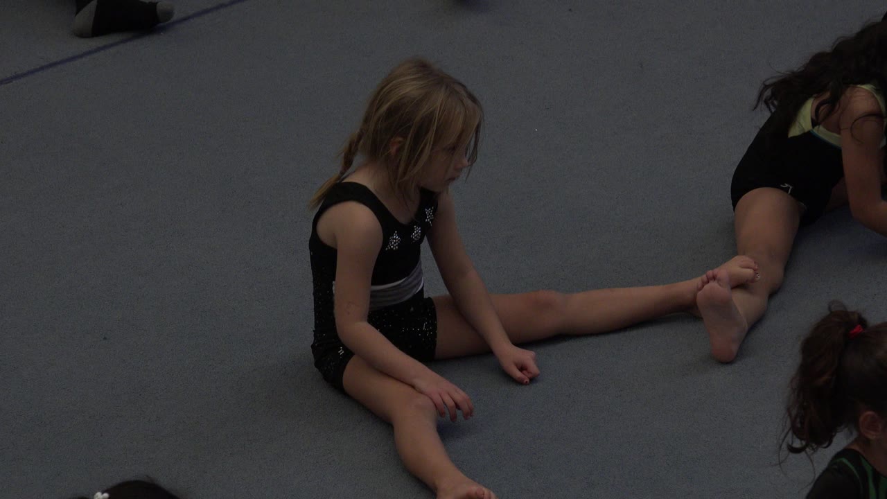 A blonde young girl sits with one leg extended on the gray floor while another child in a dark leotard sits nearby in Cabo San Lucas, Mexico.