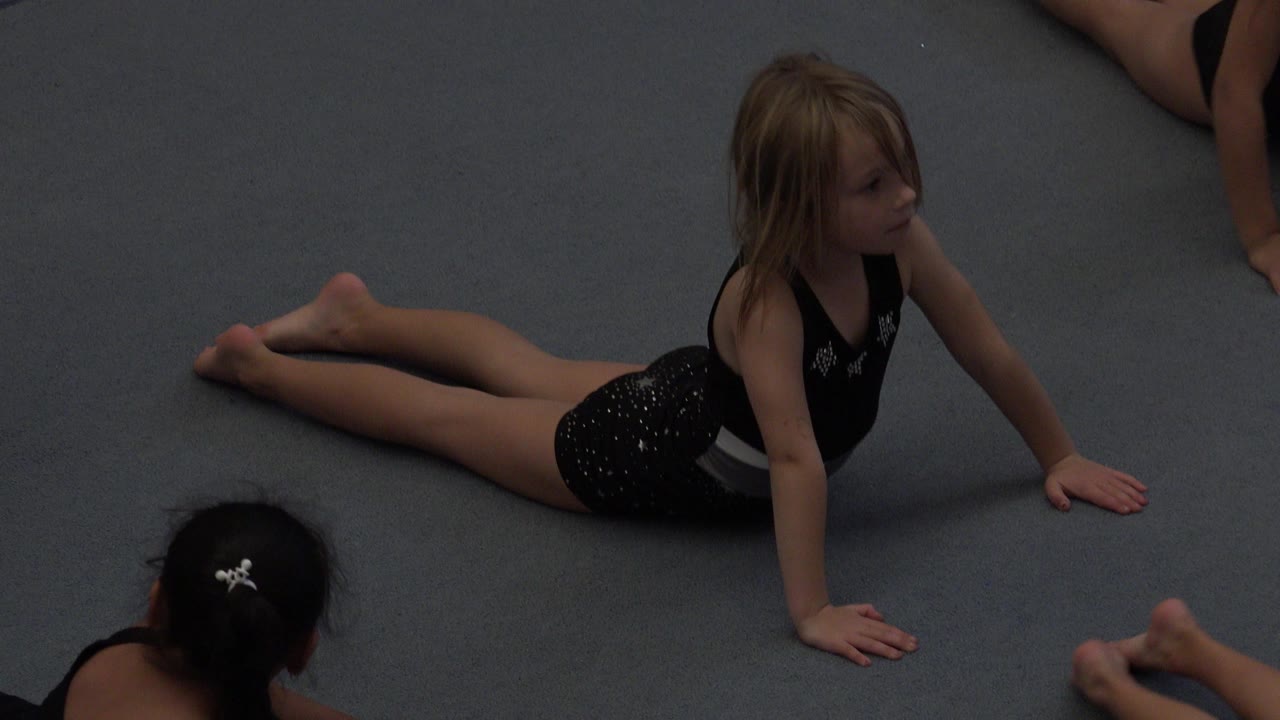 The child remains stretched on the blue floor in Cabo San Lucas while cropped arms, legs, and another child's head frame the class setting.