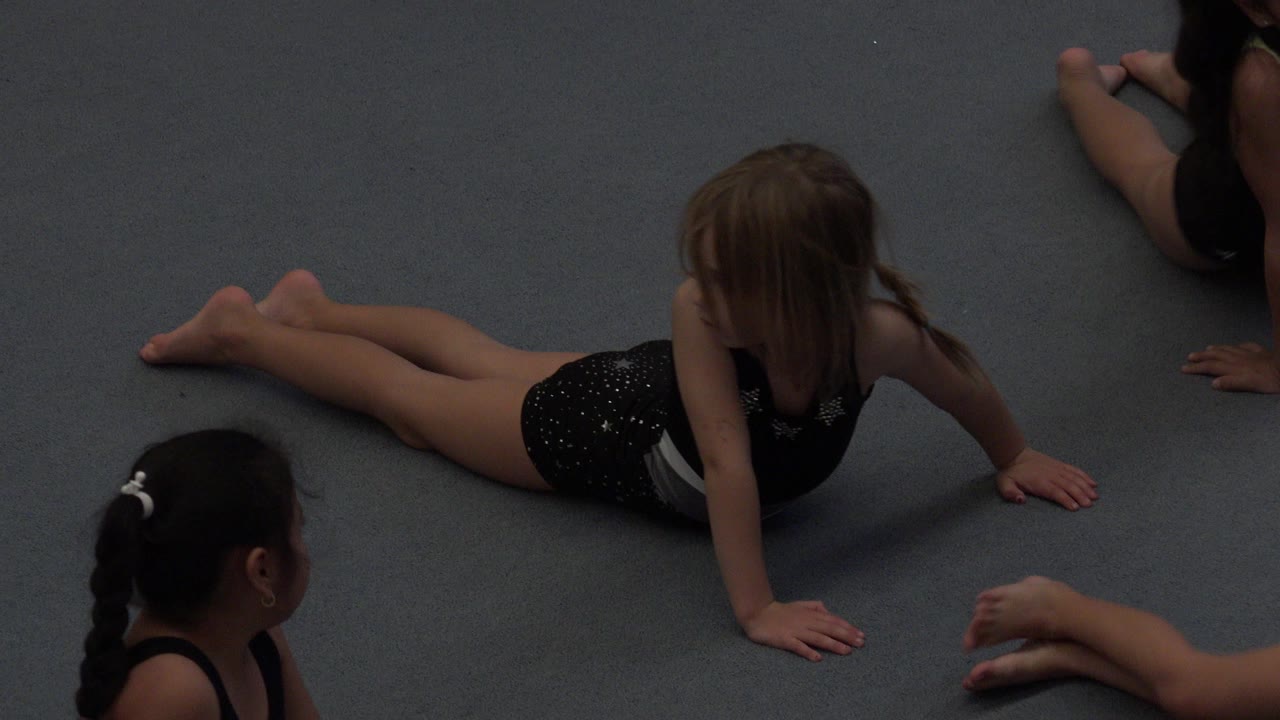 Young girl stretching on a gray gym mat with other children cropped nearby.
