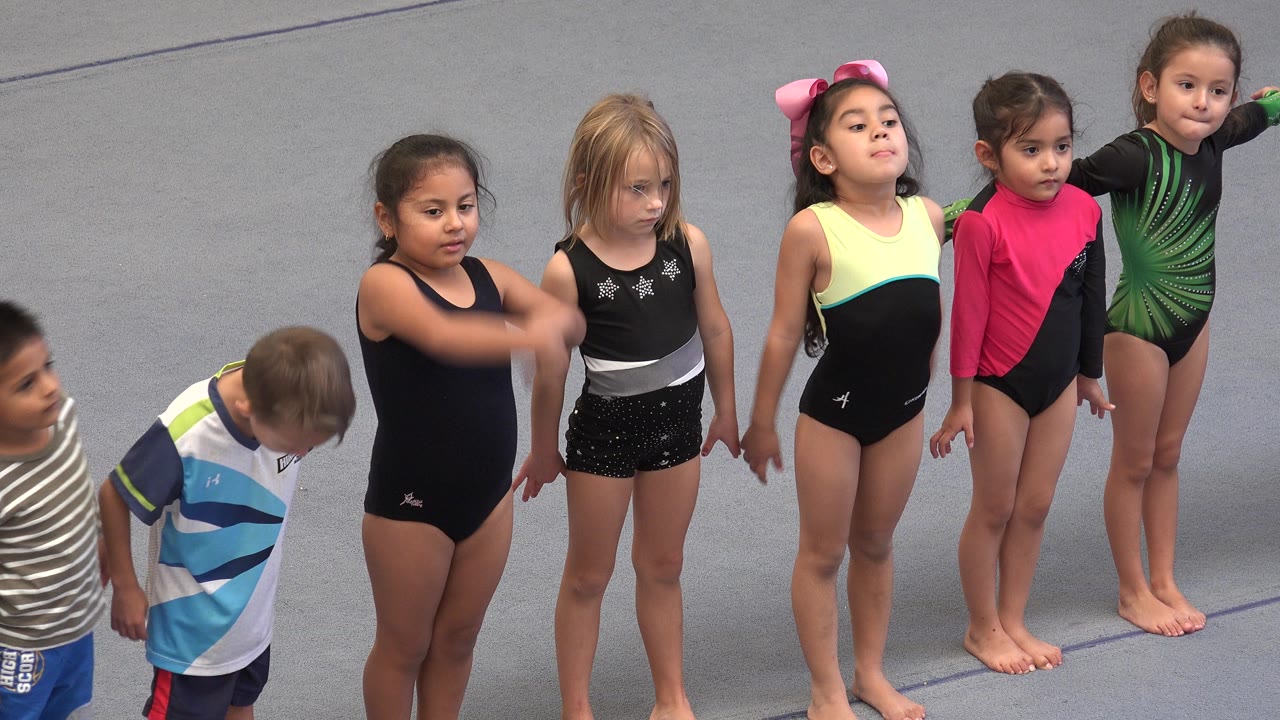 Young children stand in a loose line on a gray outdoor exercise surface in Cabo San Lucas, Mexico, with several girls in leotards and boys partly visible at the edges.