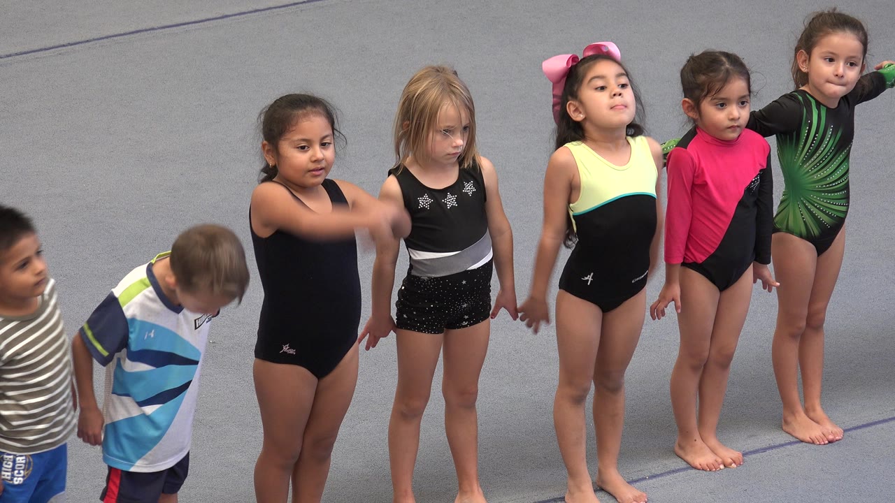 The children stay in formation on the gray floor in Cabo San Lucas, Mexico, while a girl in a black leotard starts a forward arm motion and others wait.