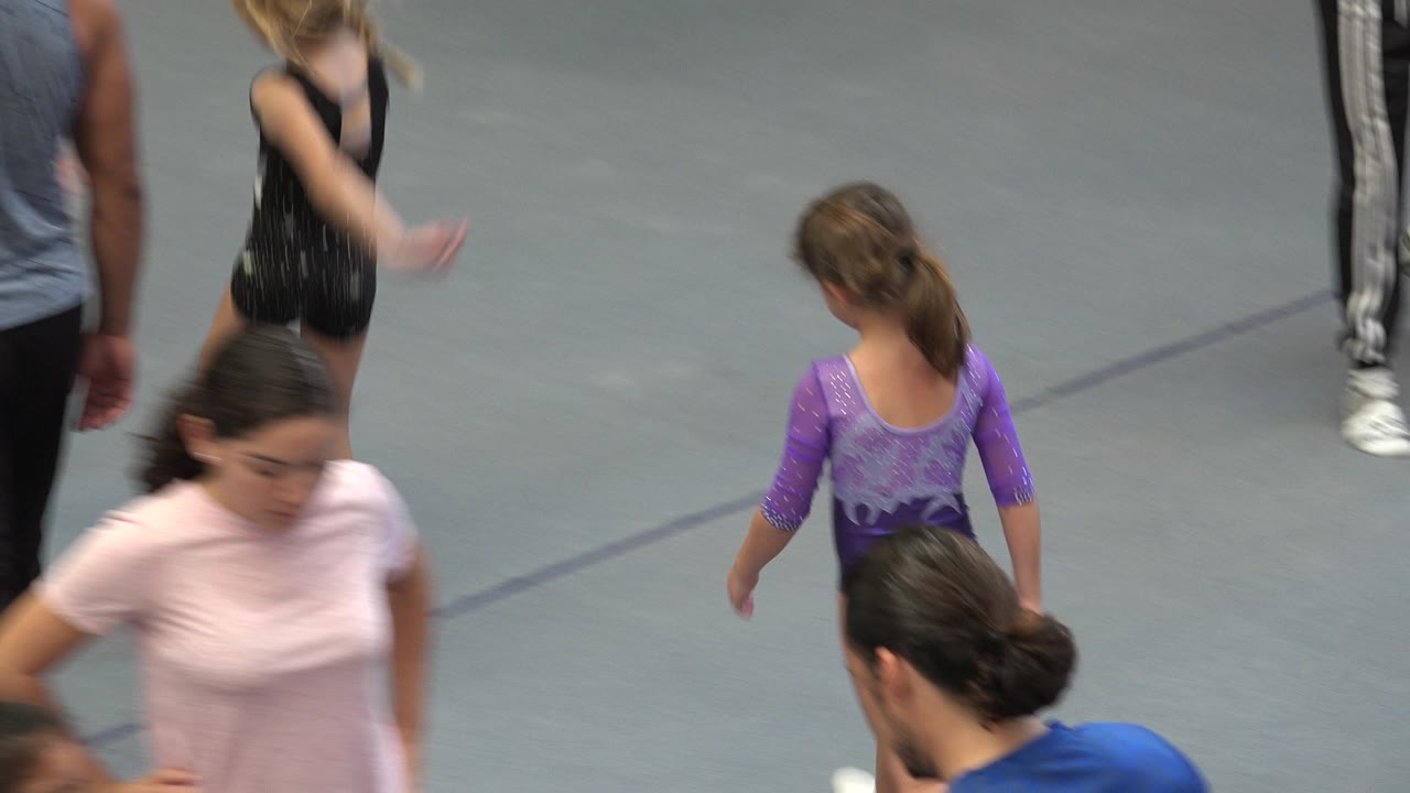 Children and adults move across a gray gymnastics floor.