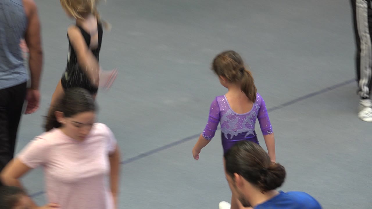 A child in a purple leotard walks away on the Cabo San Lucas gymnastics floor while adults and other children pass through the edges of the frame.