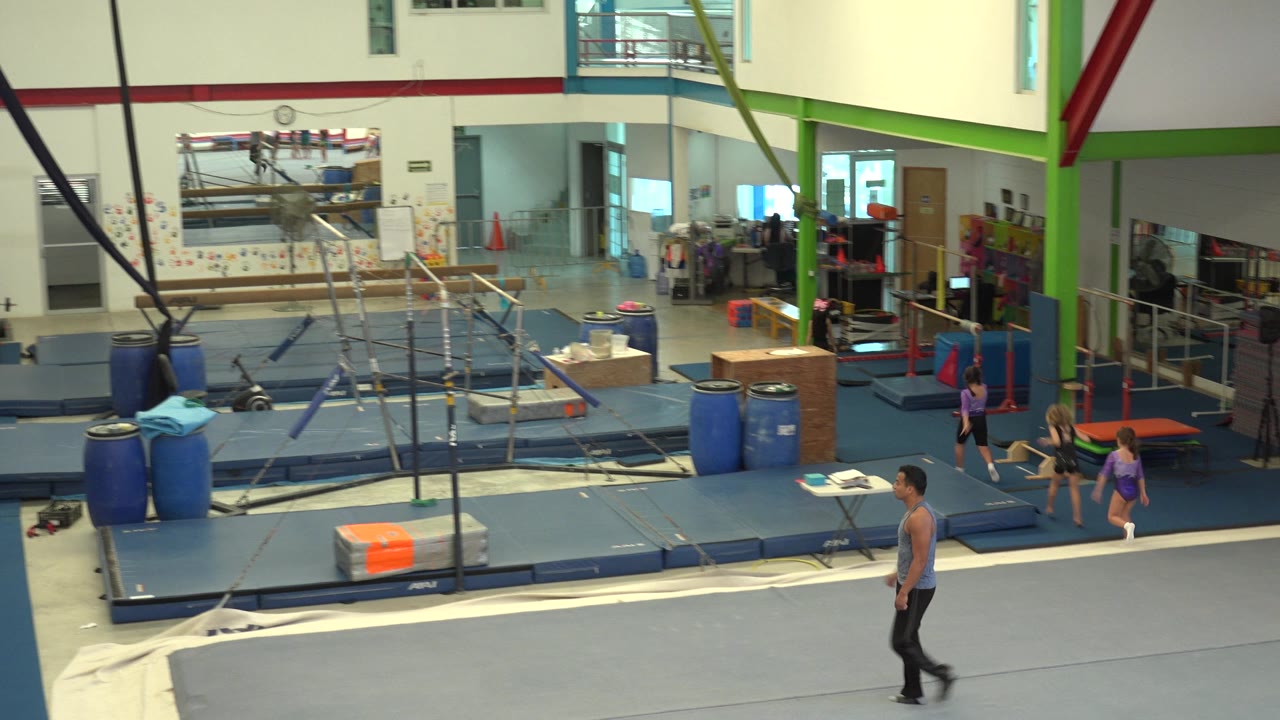 Wide view inside a Cabo San Lucas gymnasium as a man in black pants walks across a grey floor mat, with blue mats, uneven bars, ropes, and other people visible behind him.