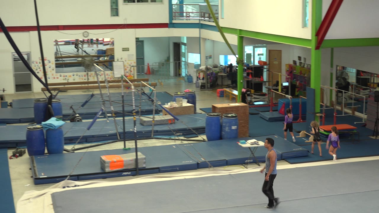 The man continues across the grey mat in the Cabo San Lucas gymnasium, with blue padded flooring, metal gymnastics bars, hanging straps, and background activity near the right wall.