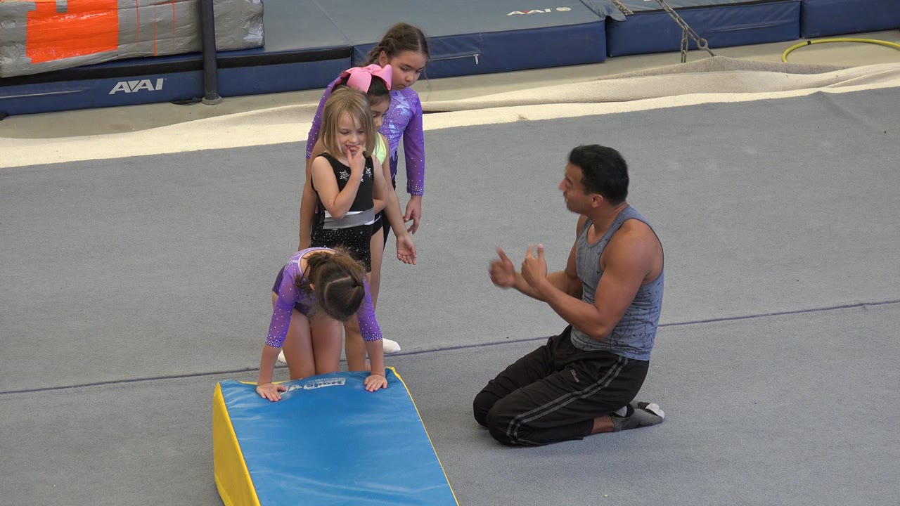 Little girls wait beside a blue wedge mat while an instructor kneels nearby.