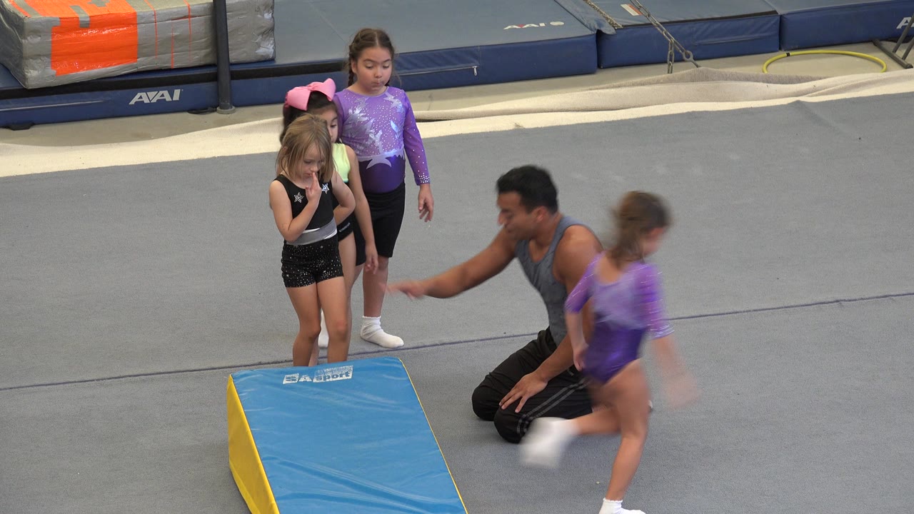 In Cabo San Lucas, young girls in leotards stand near a blue wedge mat while an adult instructor kneels on the gray gym floor and a child moves past in the foreground.