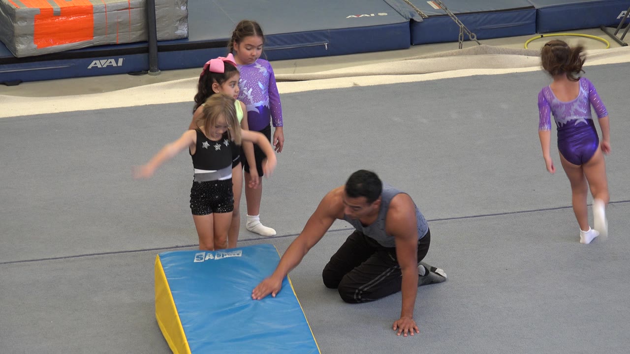 Instructor kneels near the mat as girls reposition after a turn.