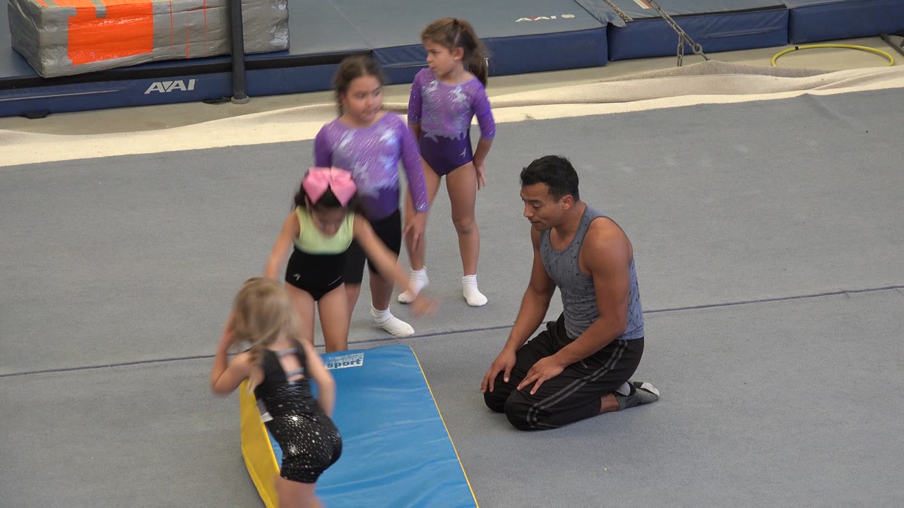 Young gymnasts stand beside a blue wedge mat while an adult coach kneels nearby in an indoor gymnastics facility in Cabo San Lucas, Mexico.