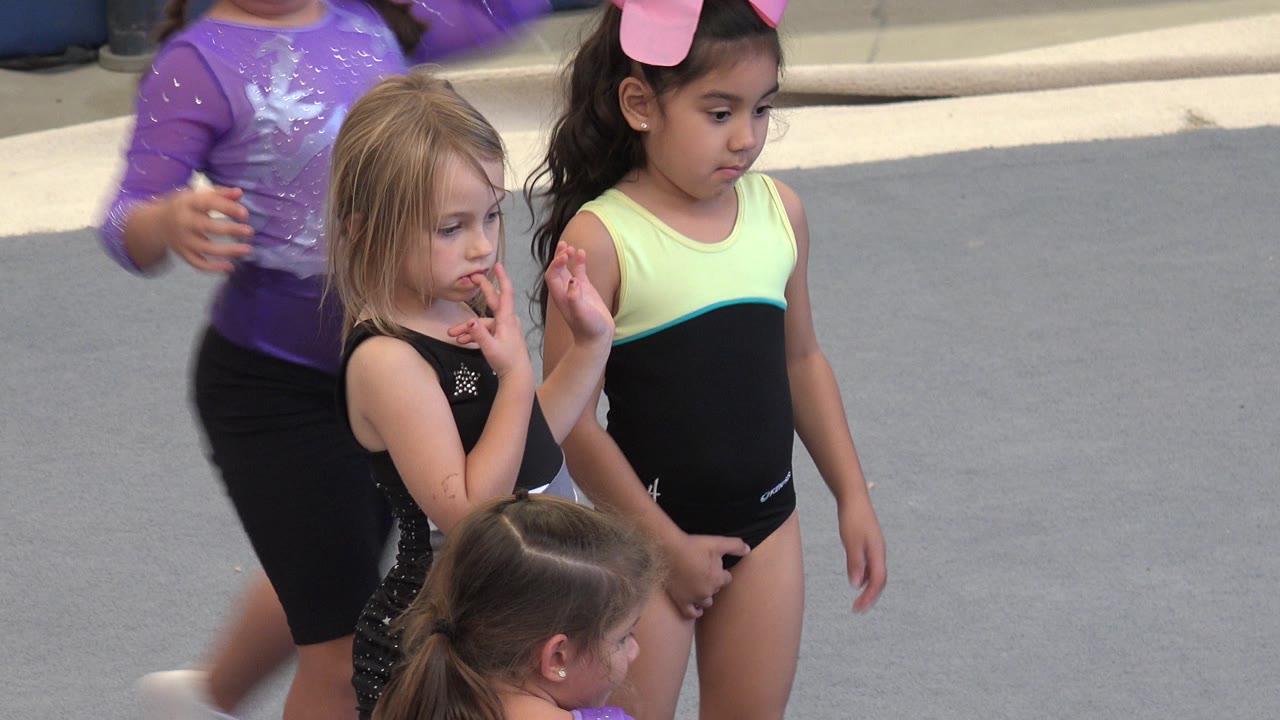Two young girls in black leotards stand in the foreground while a child in a purple leotard passes behind them on the Cabo San Lucas gym floor.