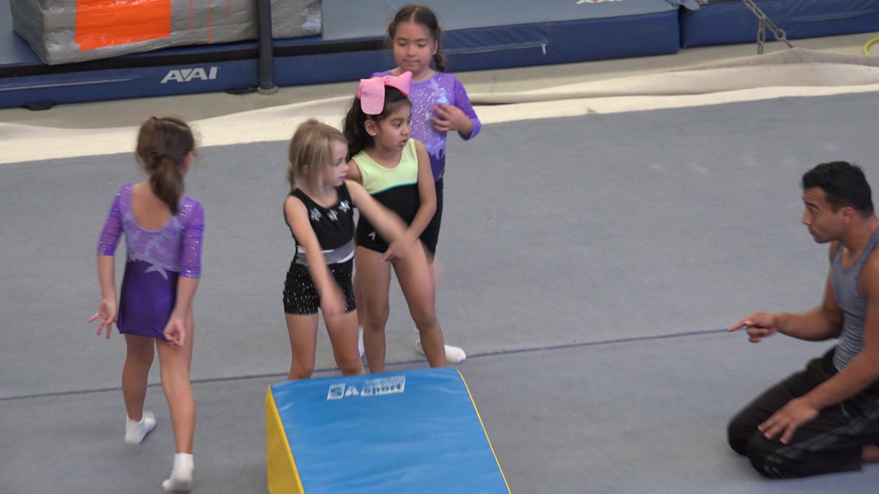 Young girls in leotards stand on a gray gym floor in Cabo San Lucas, Mexico, while a kneeling adult instructor gestures near a blue wedge mat.