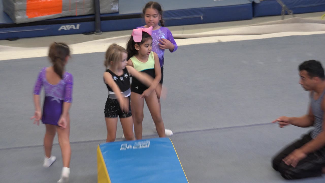 A girl in a purple leotard steps near the mat as two other girls wait in line and the adult instructor points from the right side of the Cabo San Lucas gym scene.