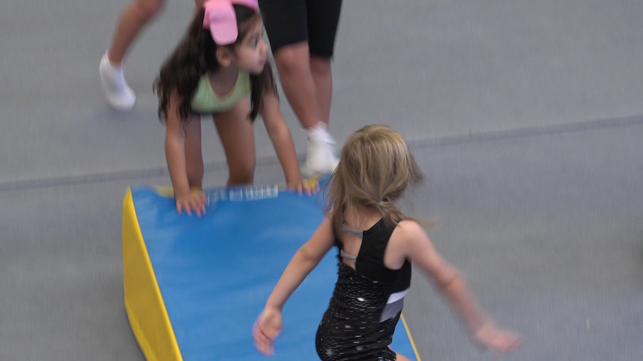 Two young children beside a blue gymnastics mat on a gray gym floor.