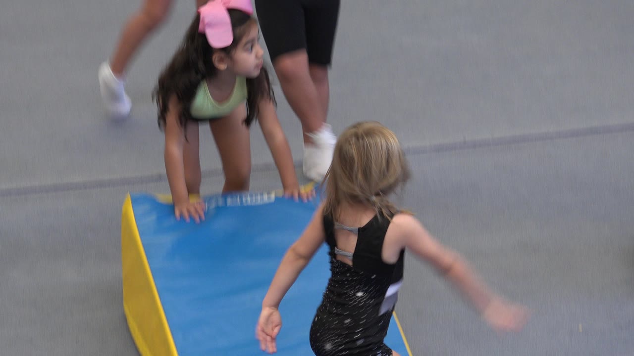 A dark-haired child leans on the blue mat while a blonde child stands in the foreground at a children's gymnastics class in Cabo San Lucas, Mexico.