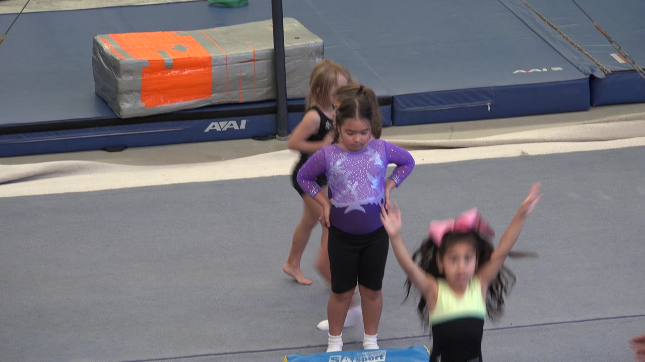 Young gymnasts stand near a blue tumbling mat as another child jumps in front.