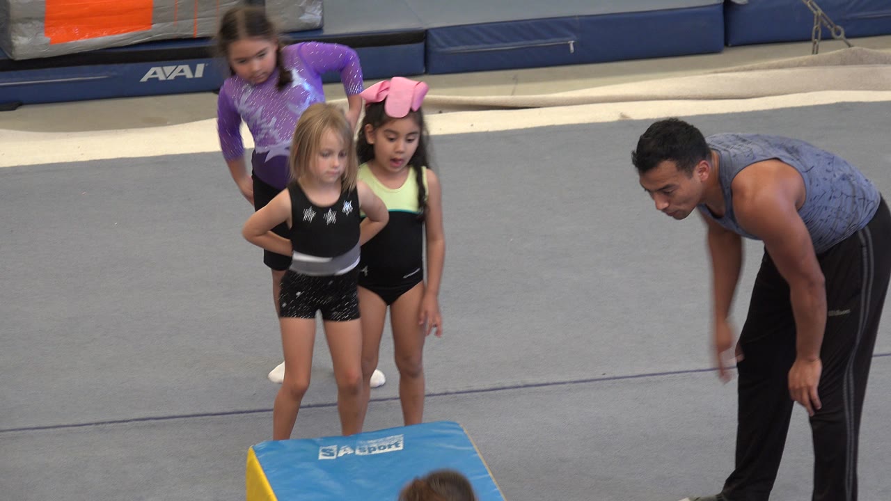 In Cabo San Lucas, young girls in leotards stand near a blue padded gymnastics block while a male instructor bends forward on the gray gym floor.