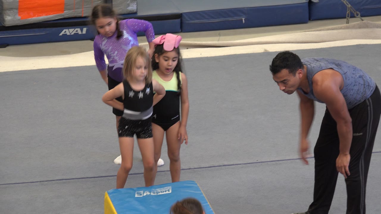 In Cabo San Lucas, the instructor leans toward the blue gymnastics block while three girls wait close together on the indoor gym floor.
