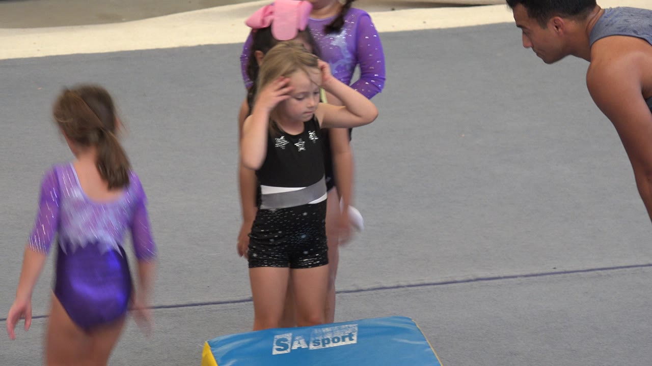 Young girl in black leotard beside a blue gymnastics mat with coach at right.