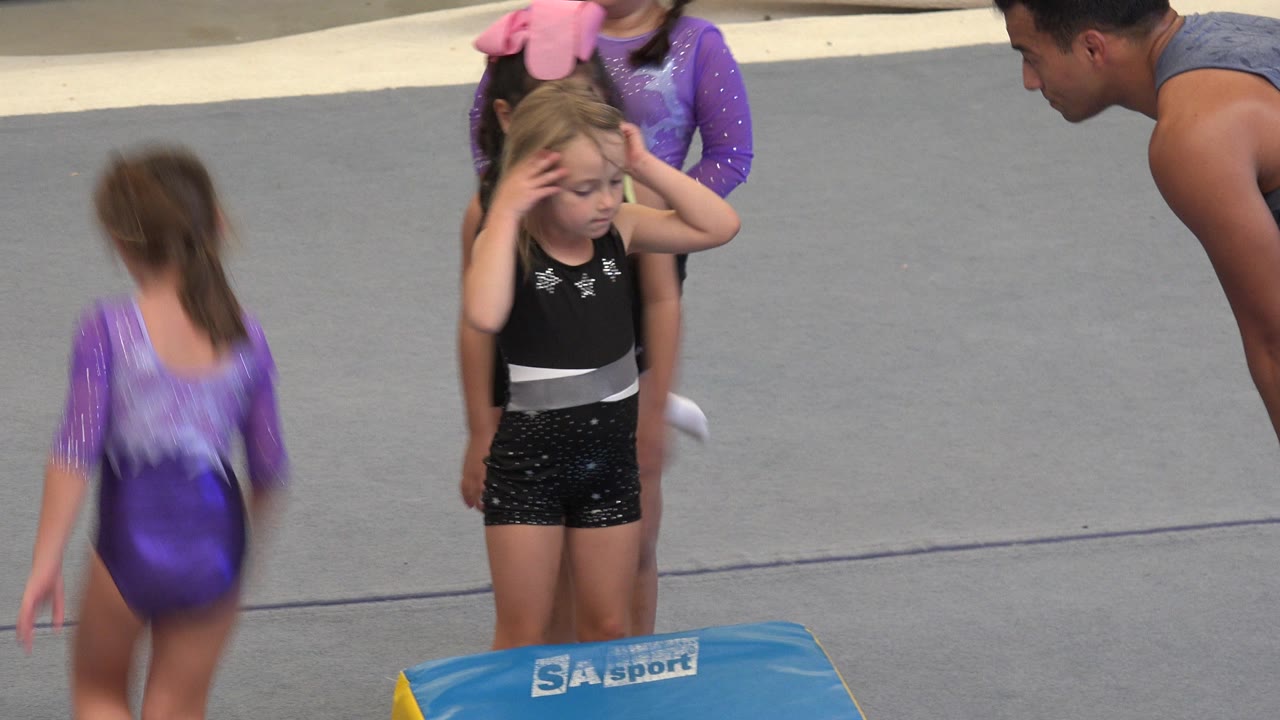 In Cabo San Lucas, Mexico, the young girl remains beside the blue mat with her hands near her face as a child in a purple leotard moves across the foreground and the coach watches from the right.