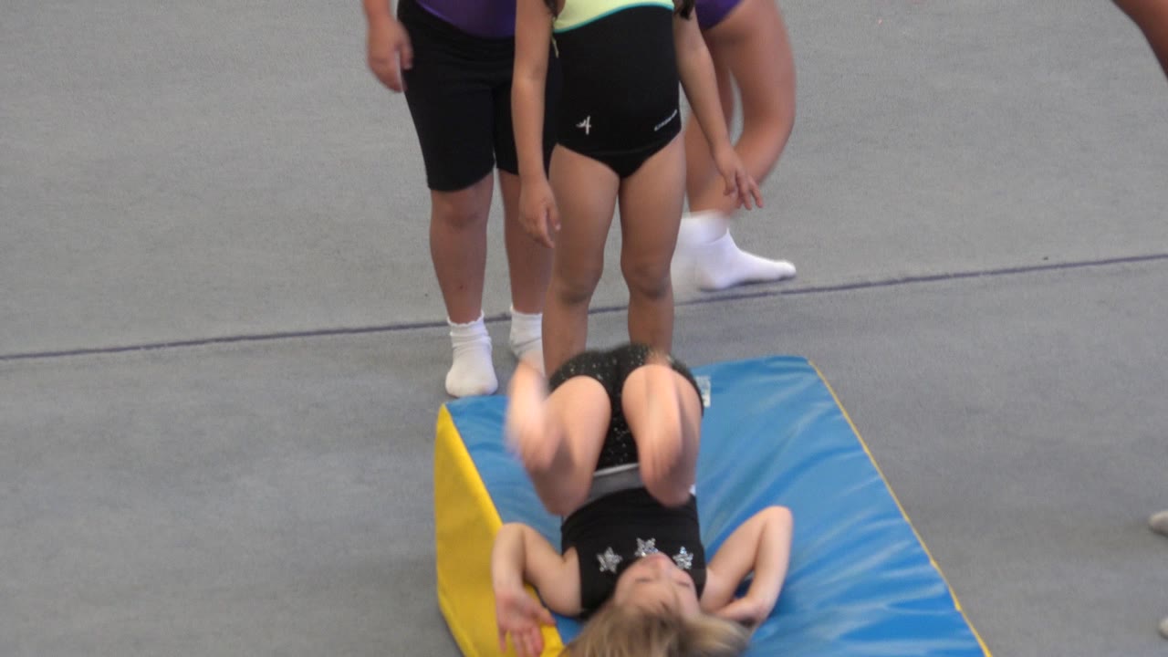 Girl lying back on a blue gym mat while adults stand nearby.