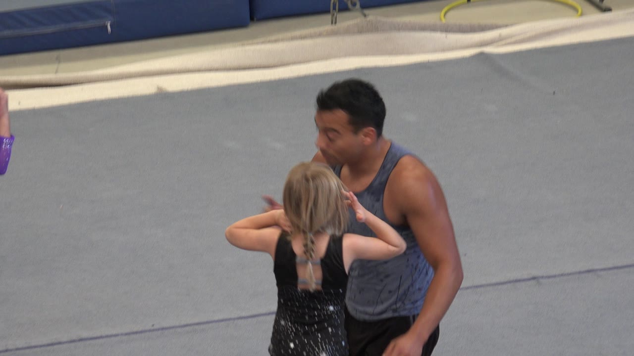 Coach kneeling beside a young girl in a black dance outfit on a gray mat.