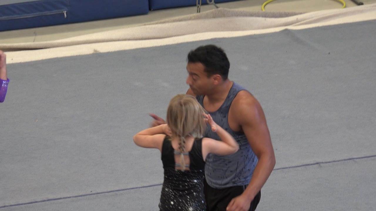 In Cabo San Lucas, the coach stays kneeling beside the blonde child in black while the dancer holds her hands near her head on the gray mat.