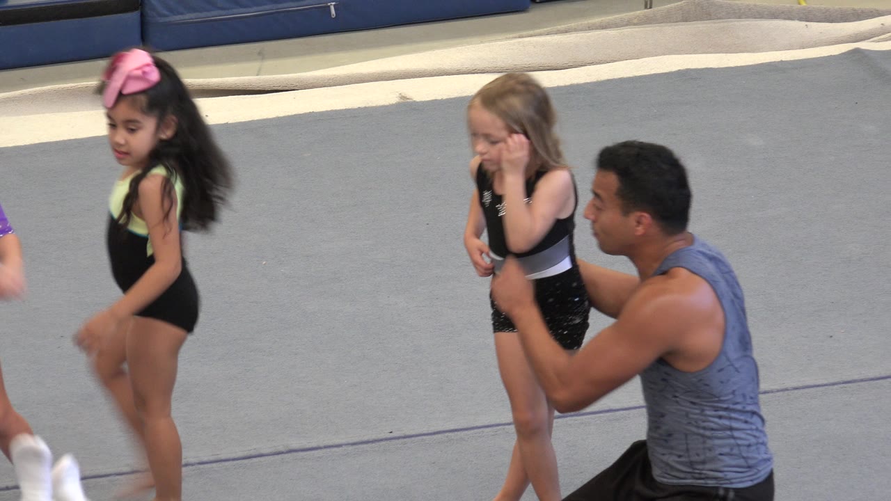 At 0:00 in Cabo San Lucas, young girls in leotards stand on gray gymnastics mats while a male instructor kneels beside one child and blue padding lines the background.